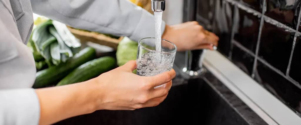 A person filling a glass with water from a kitchen faucet next to fresh vegetables.