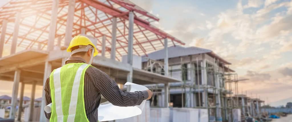 A construction site with a steel frame building and a worker in a yellow hard hat reviewing blueprints.