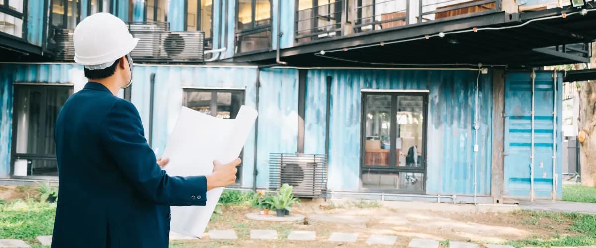 Engineer in a white hard hat inspecting a modern container home with architectural blueprints in hand.