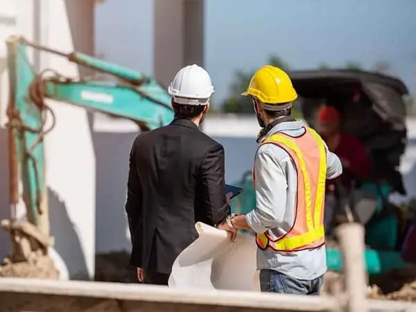 Construction site discussion between a project manager in a suit and a worker in a safety vest and hard hat, holding blueprints